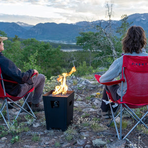The Live Outdoor Firestorm Series I Free Standing Fire Pit is being used outdoors, providing a great view of the mountains, with people sitting nearby.