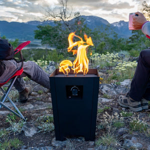 The Live Outdoor Firestorm Series I Free Standing Fire Pit is being used outdoors with people sitting nearby.