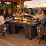 Friends passing dishes at a backyard dinner around a Top Fires Alameda 72-inch GFRC gas fire table with a Metallic Slate tabletop and Copper Vein base