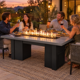 Couples toasting at a dinner party around a Top Fires Alameda 72-inch GFRC gas fire table with a Natural Gray tabletop and Black base
