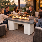 Friends passing dishes at a backyard dinner around a Top Fires Alameda 72-inch GFRC gas fire table with a Natural Gray tabletop and White base